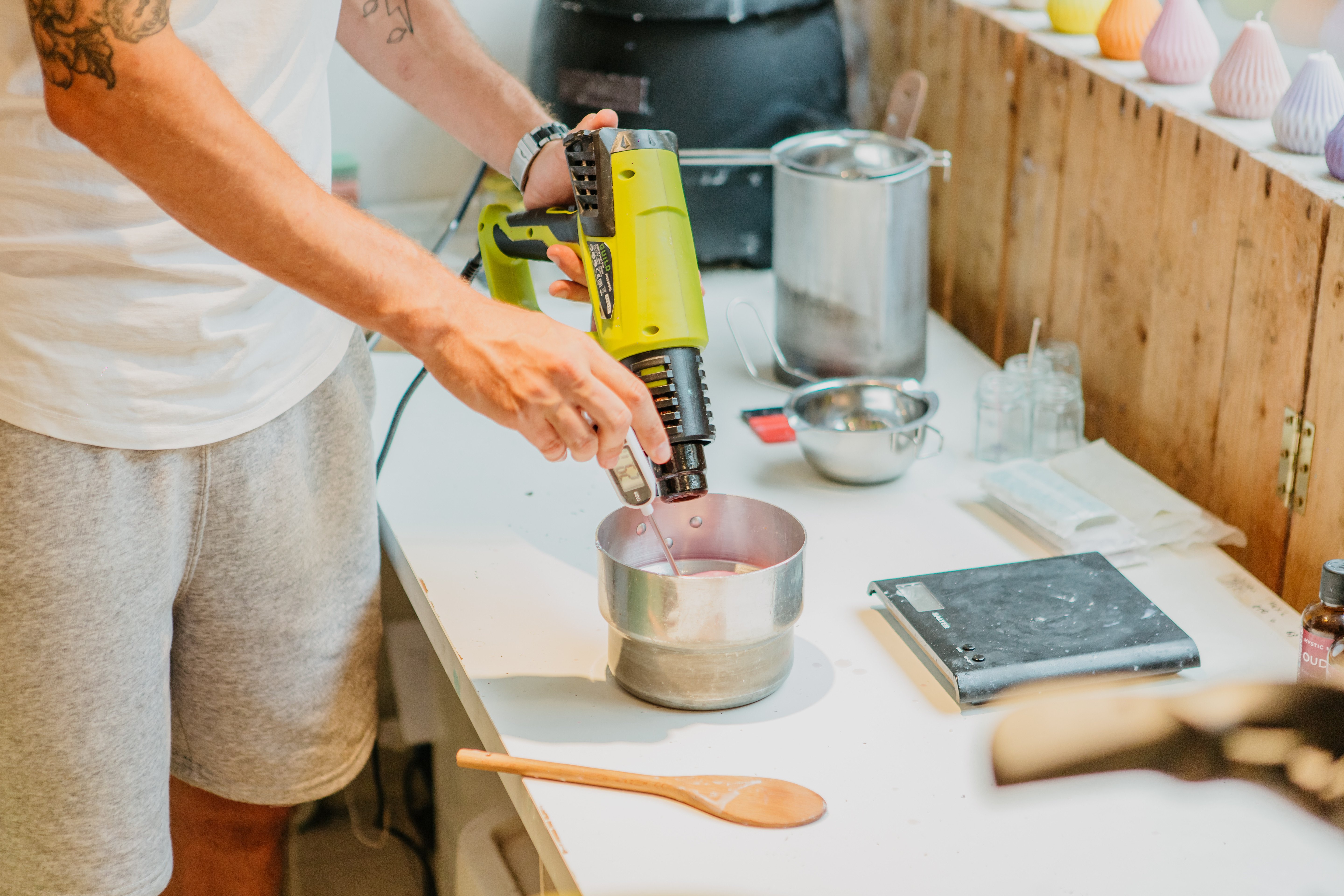 A man warms up candle wax in a metal container using a heat gun